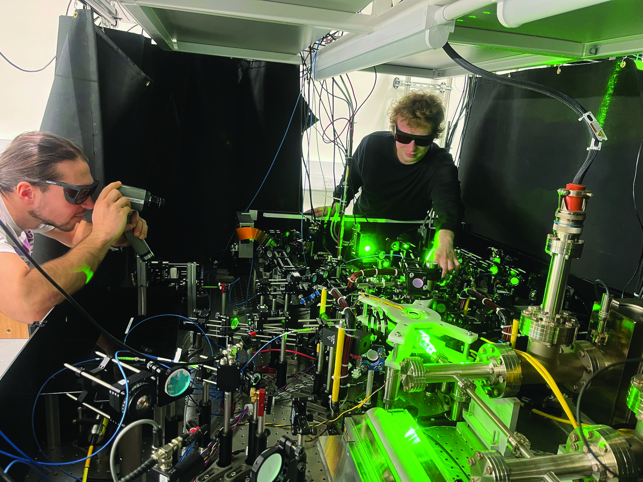Two people wearing sunglasses lean over a table covered with metal parts, wires, and green lasers. 