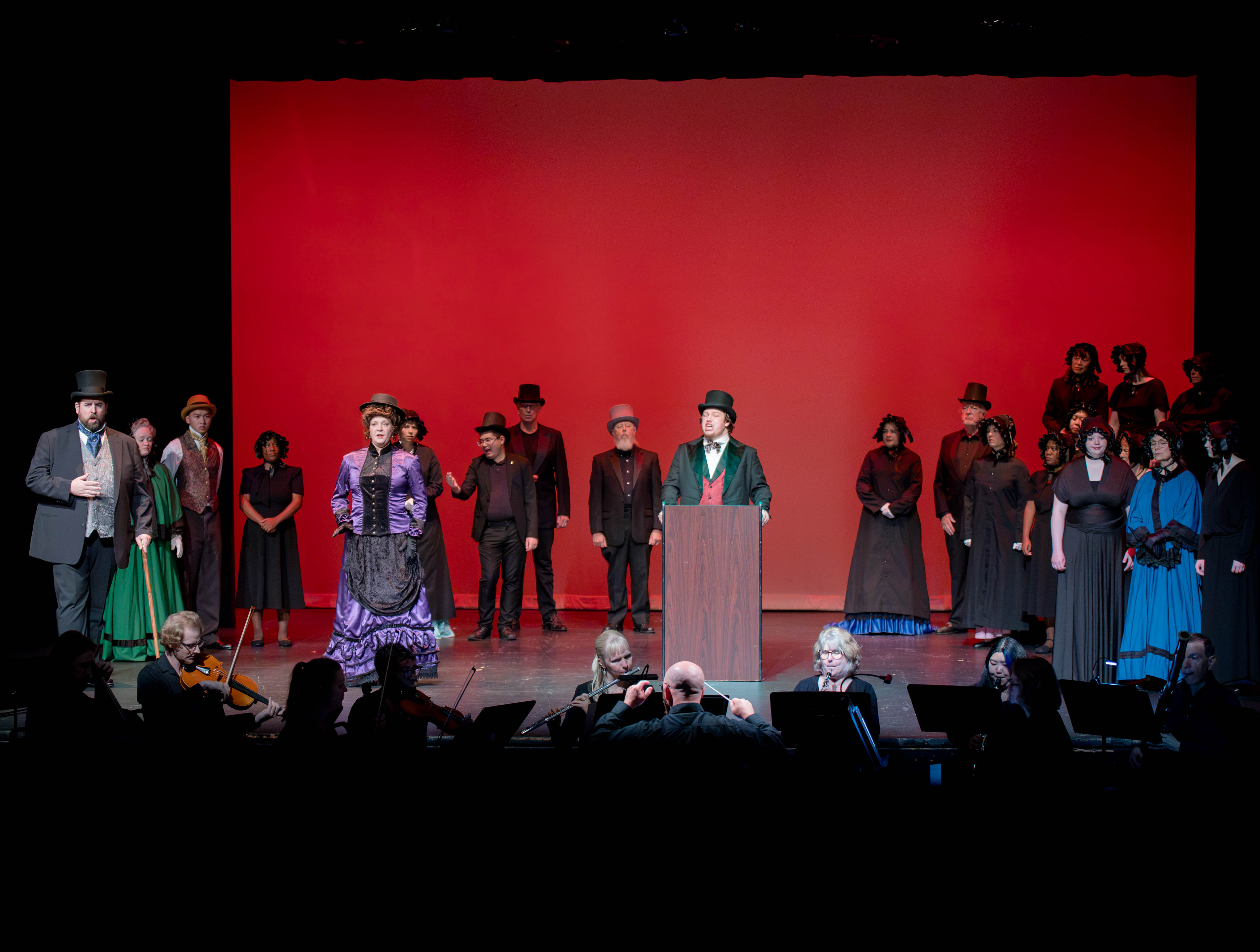 Man stands at podium with singers to the left and right. Orchestra is in foreground.