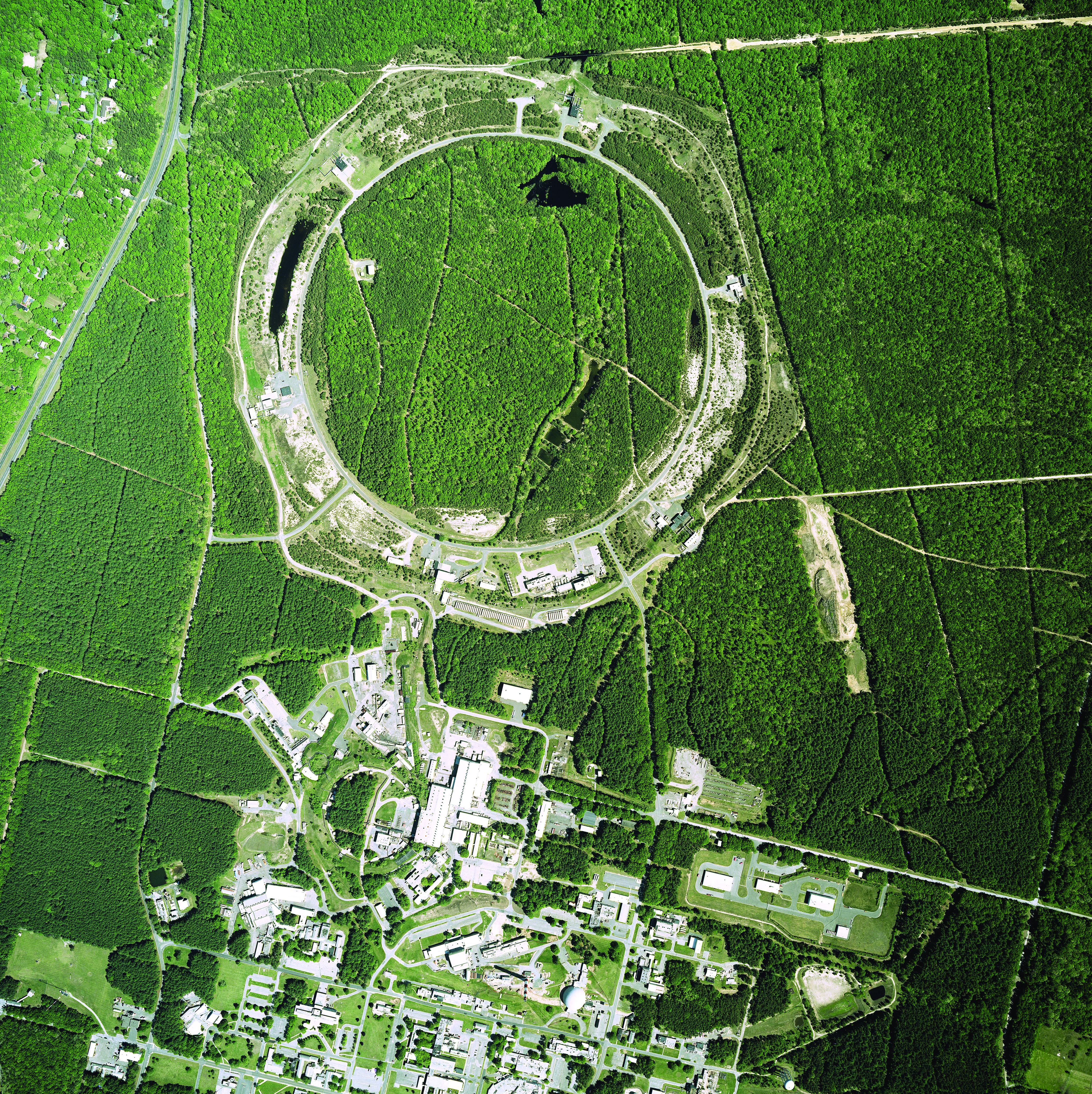 An aerial photo of green fields that host a large complex of white buildings with a large circular building at the north end. 