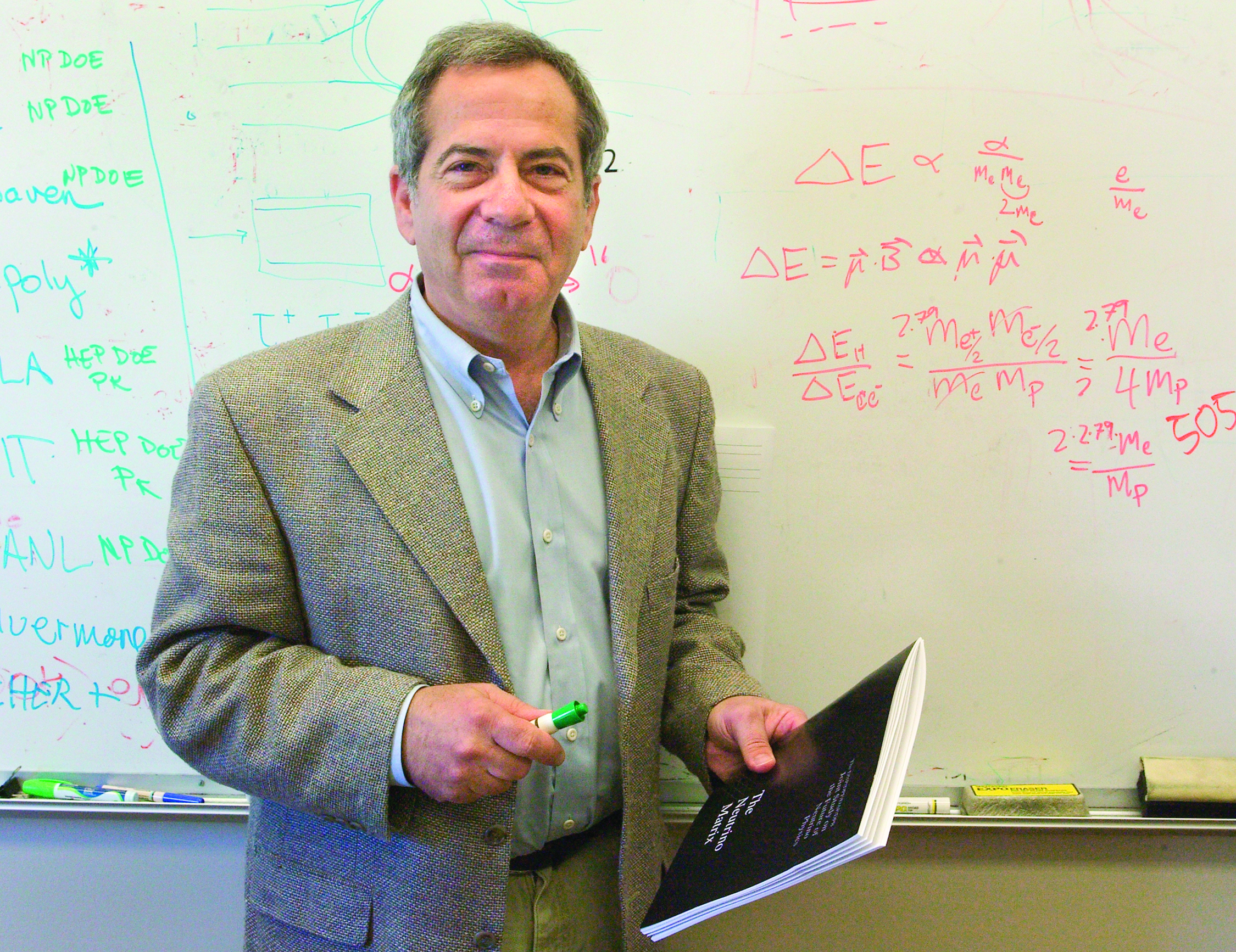 A man holding a booklet in front of a whiteboard with writing on it.