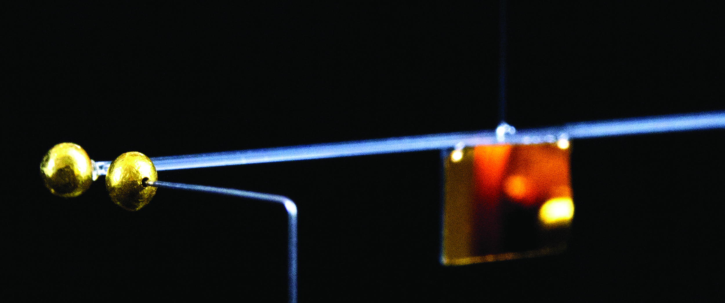 A torsion pendulum is depicted with a gold sphere on one end of a metal wire and a second, fixed gold sphere positioned nearby.