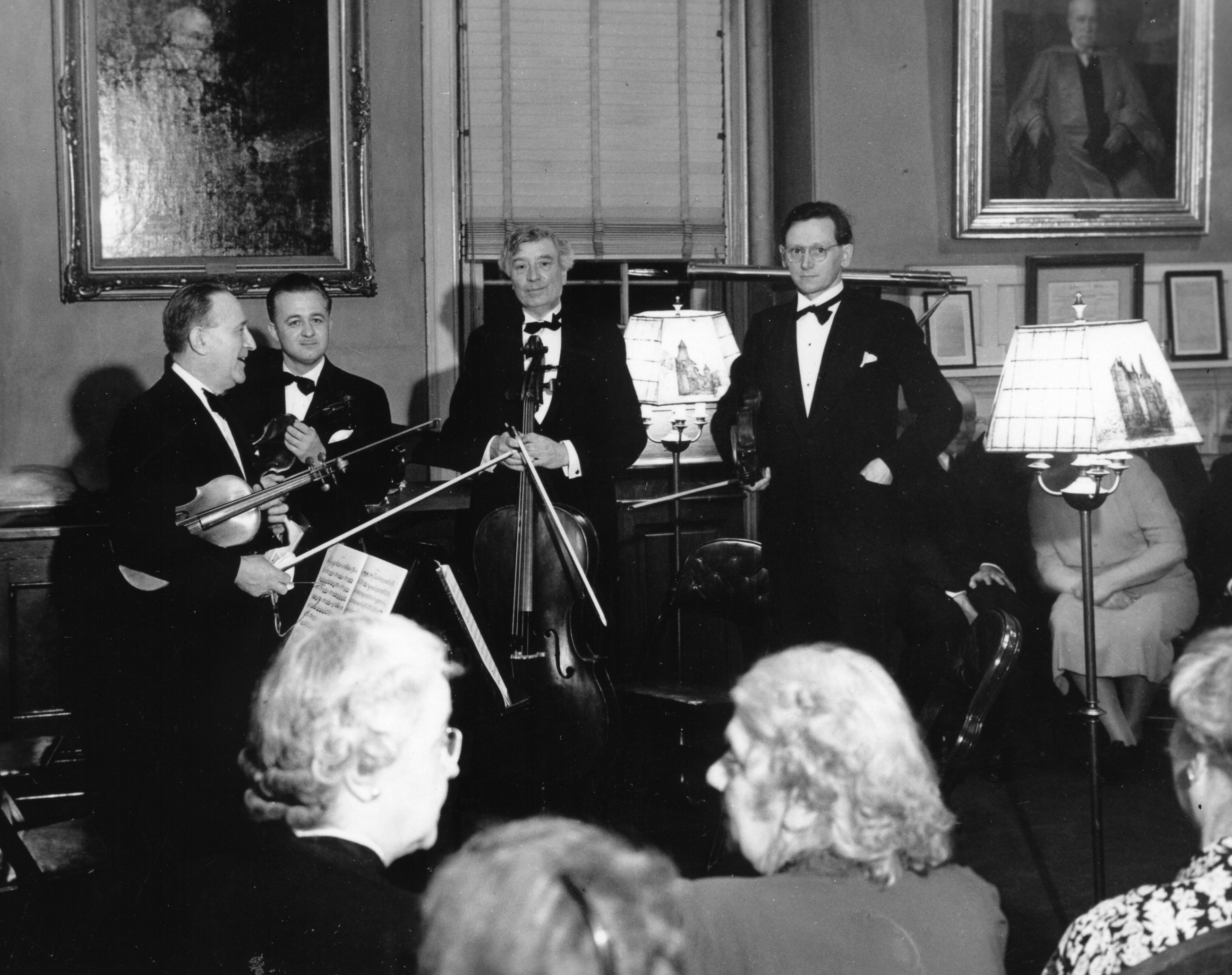 String quartet stands after a performance in a parlor with paintings on the wall, Swann is holding a cello. Grey haired women are in sitting in the foreground.