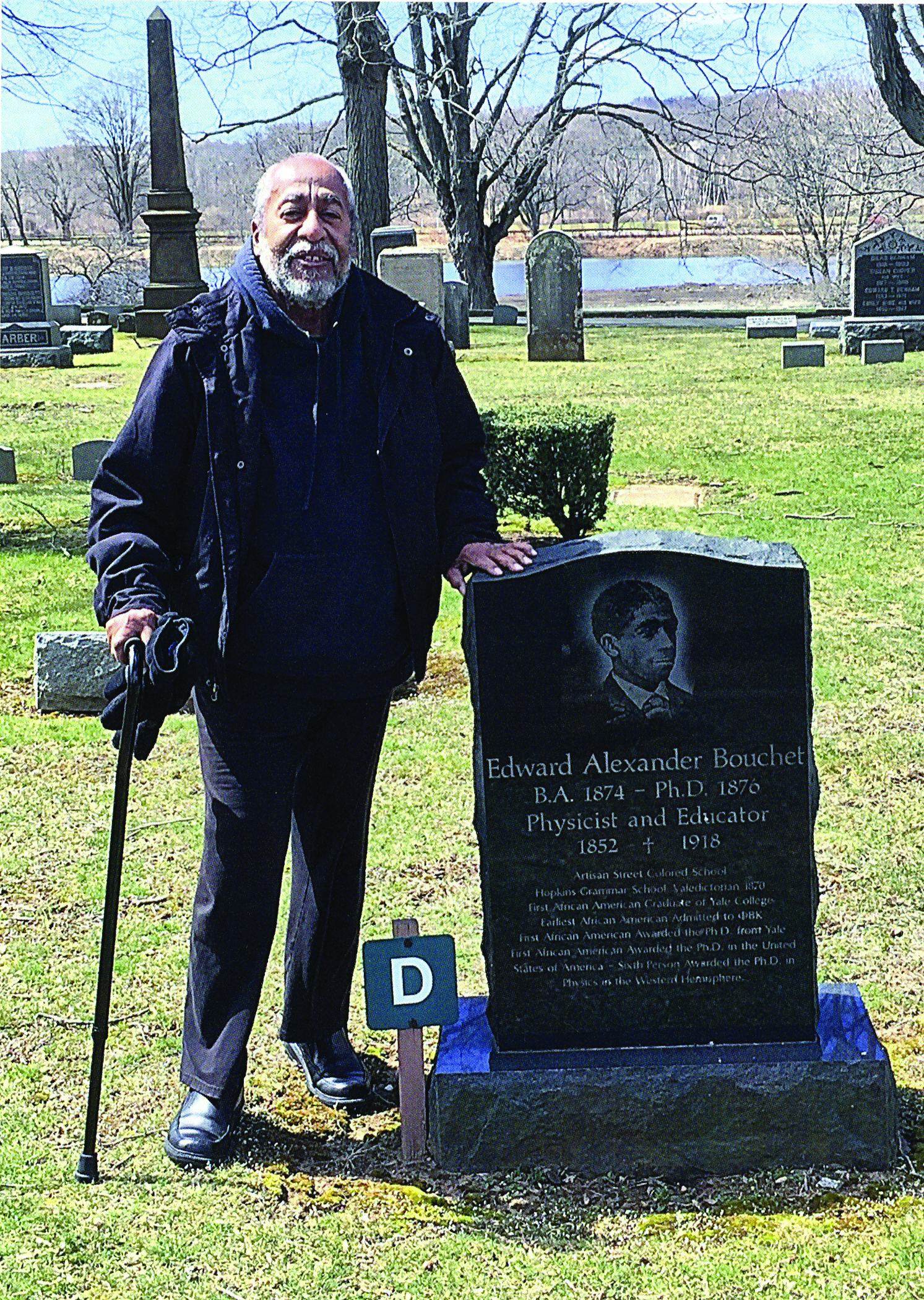 An older man with a cane stands at the side of a gravestone.