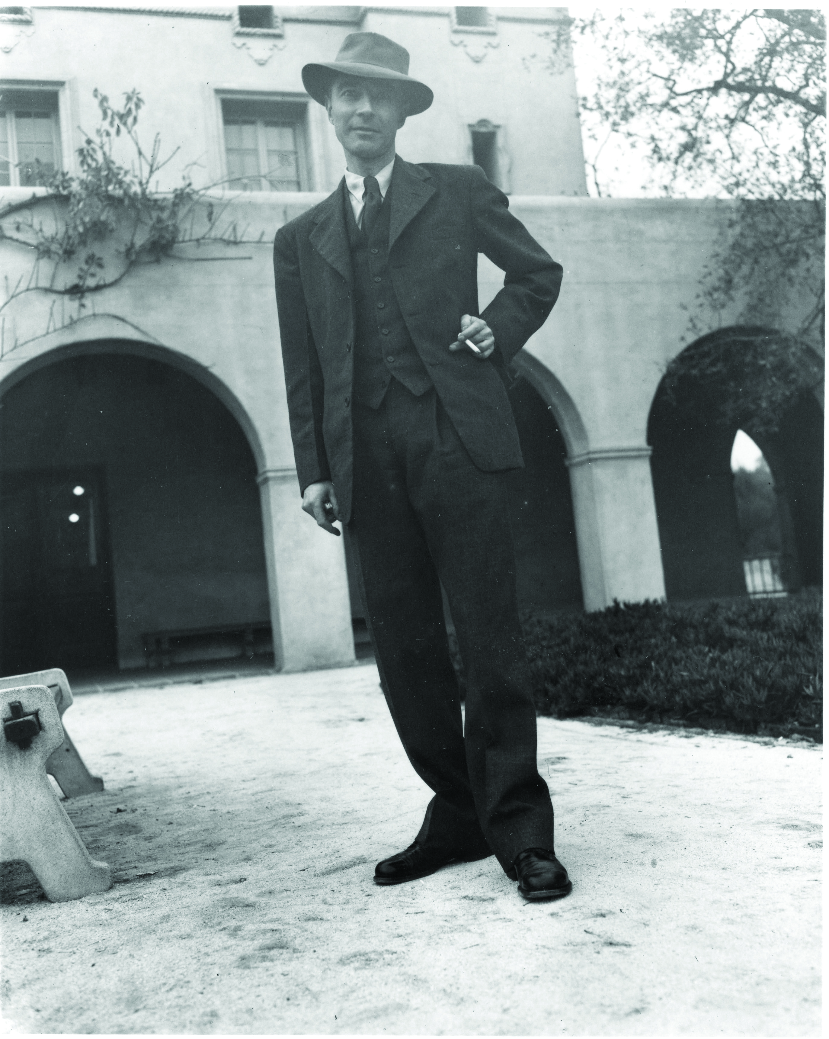 A man stands jauntily, wearing a porkpie hat and holding a cigarette.