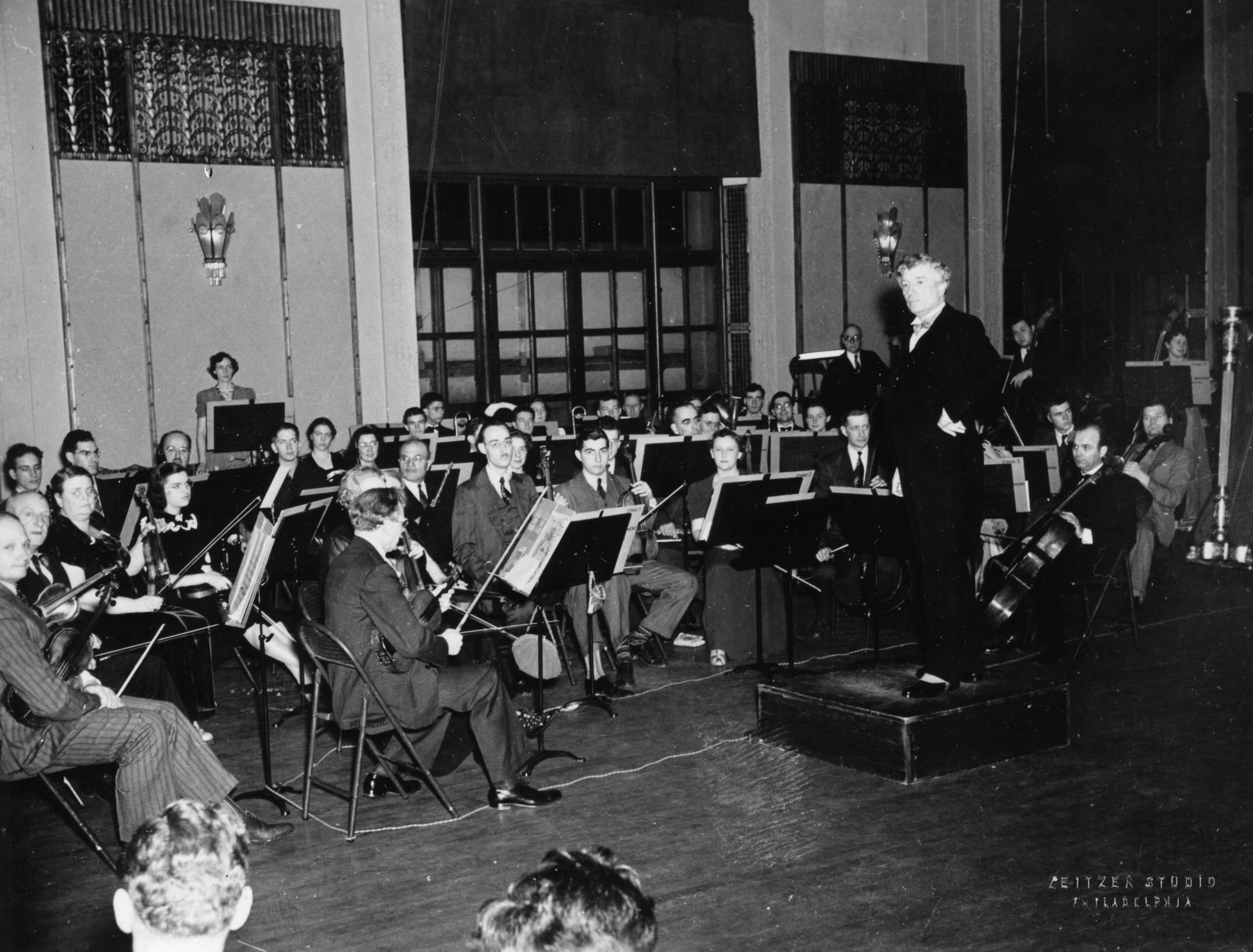 Orchestra prepared to play in a concert hall with Swann standing on a podium holding a baton.