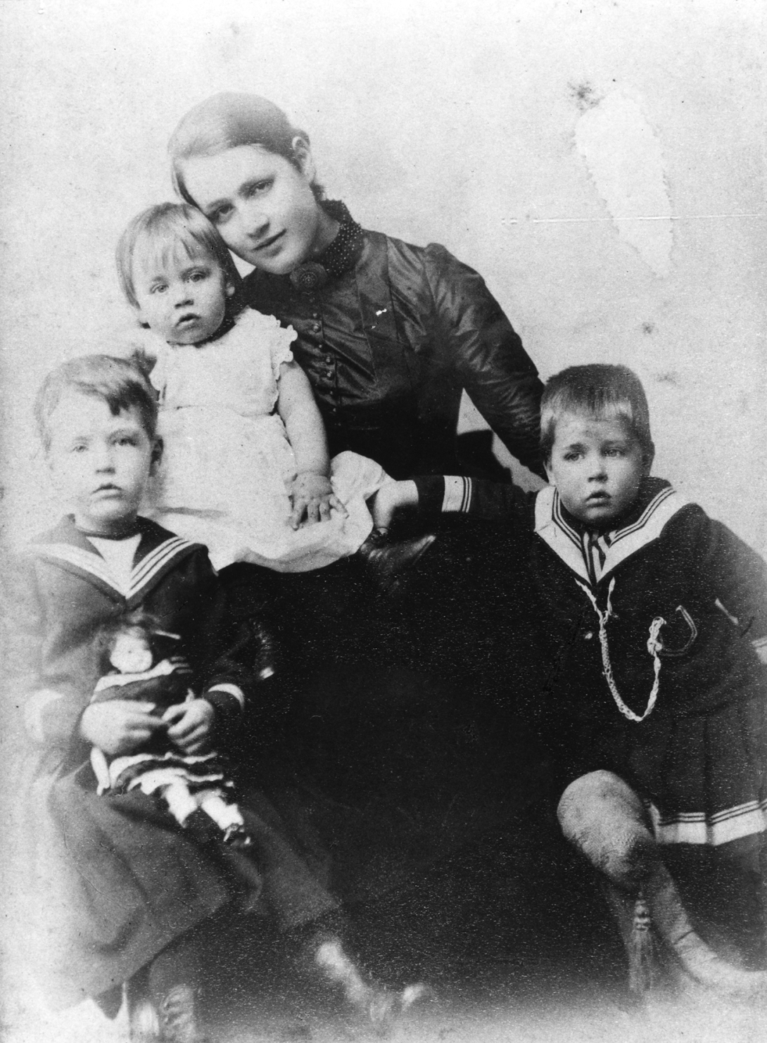 Group portrait of a seated woman with a baby girl in her lap. A boy is seated at her feat on the left and holds a doll. Another boy is leaning toward the woman on the right.