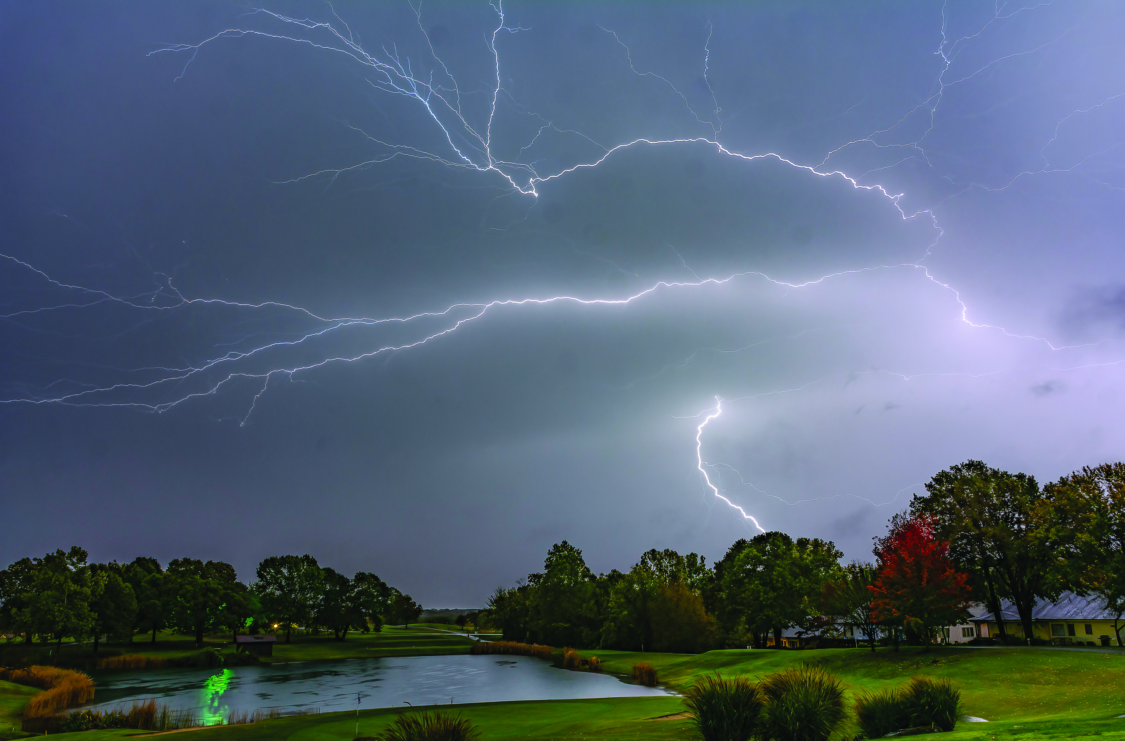Multiple lightning bolts in a sky of dark gray clouds over a green grassy area with trees and a pond.