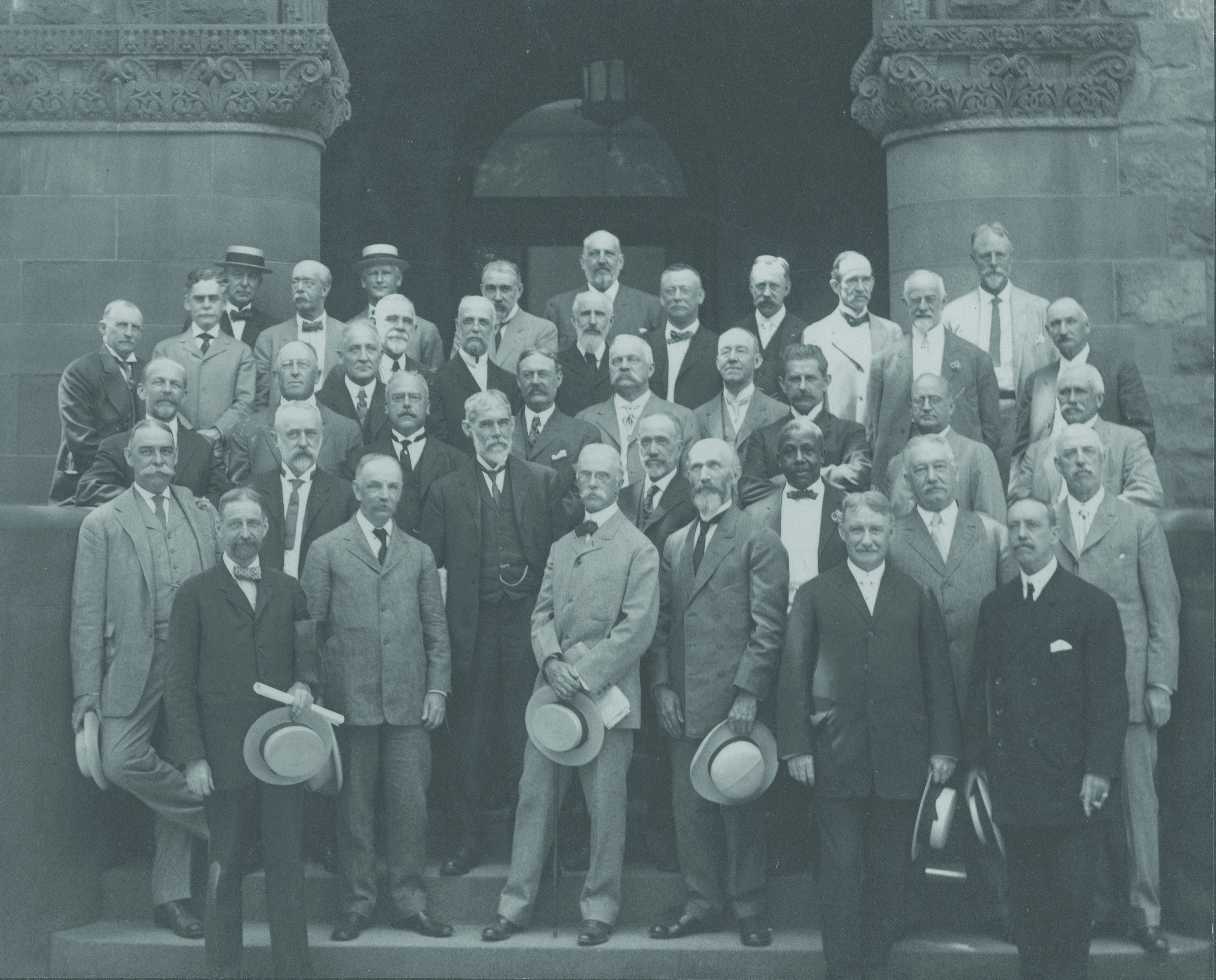 A group of older men dressed in suits and ties stand in front of a building. One of the men is Black; all the others are white.