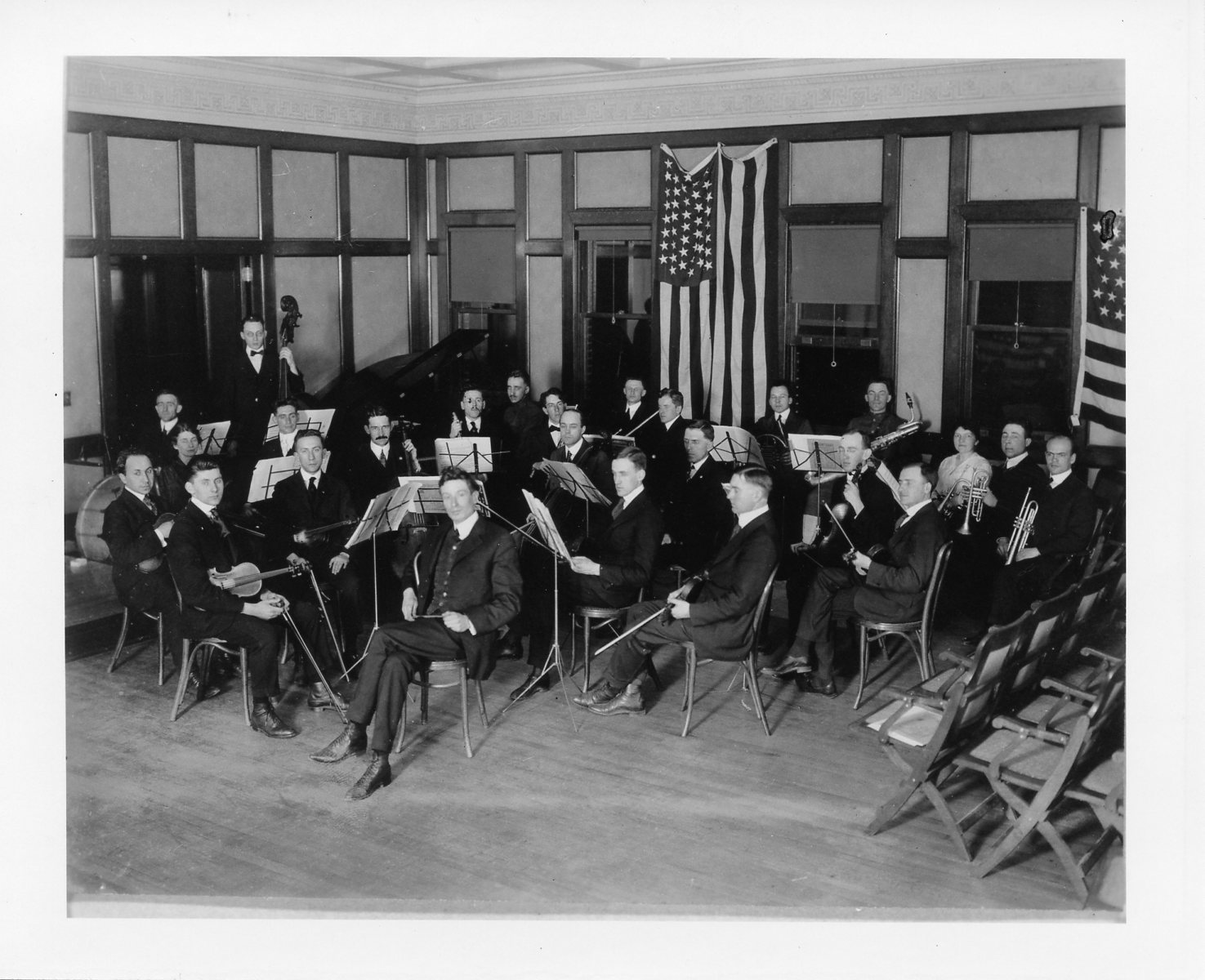 Conductor seated in front of an orchestra in a room with American flags in the background.