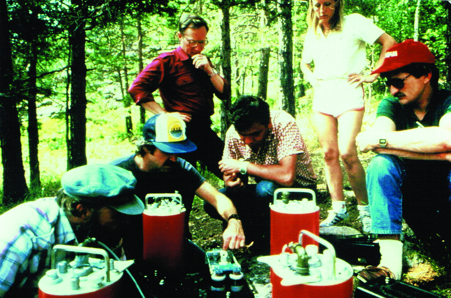 Four men sit and two stand around a collection of red cylinders with protrusions and handles on top.