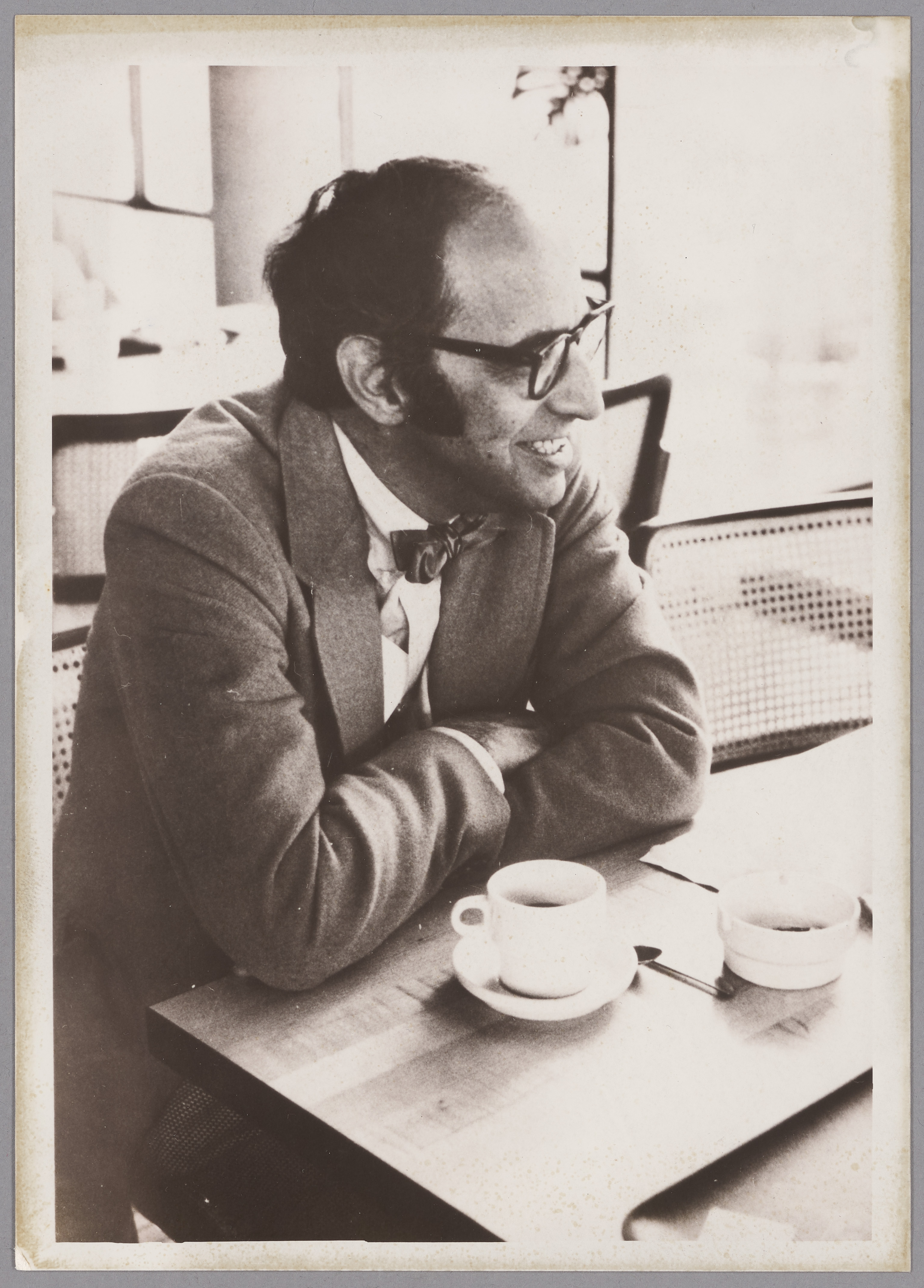 A middle-aged man wearing a suit and bow tie smiles with a cup of coffee in front of him.