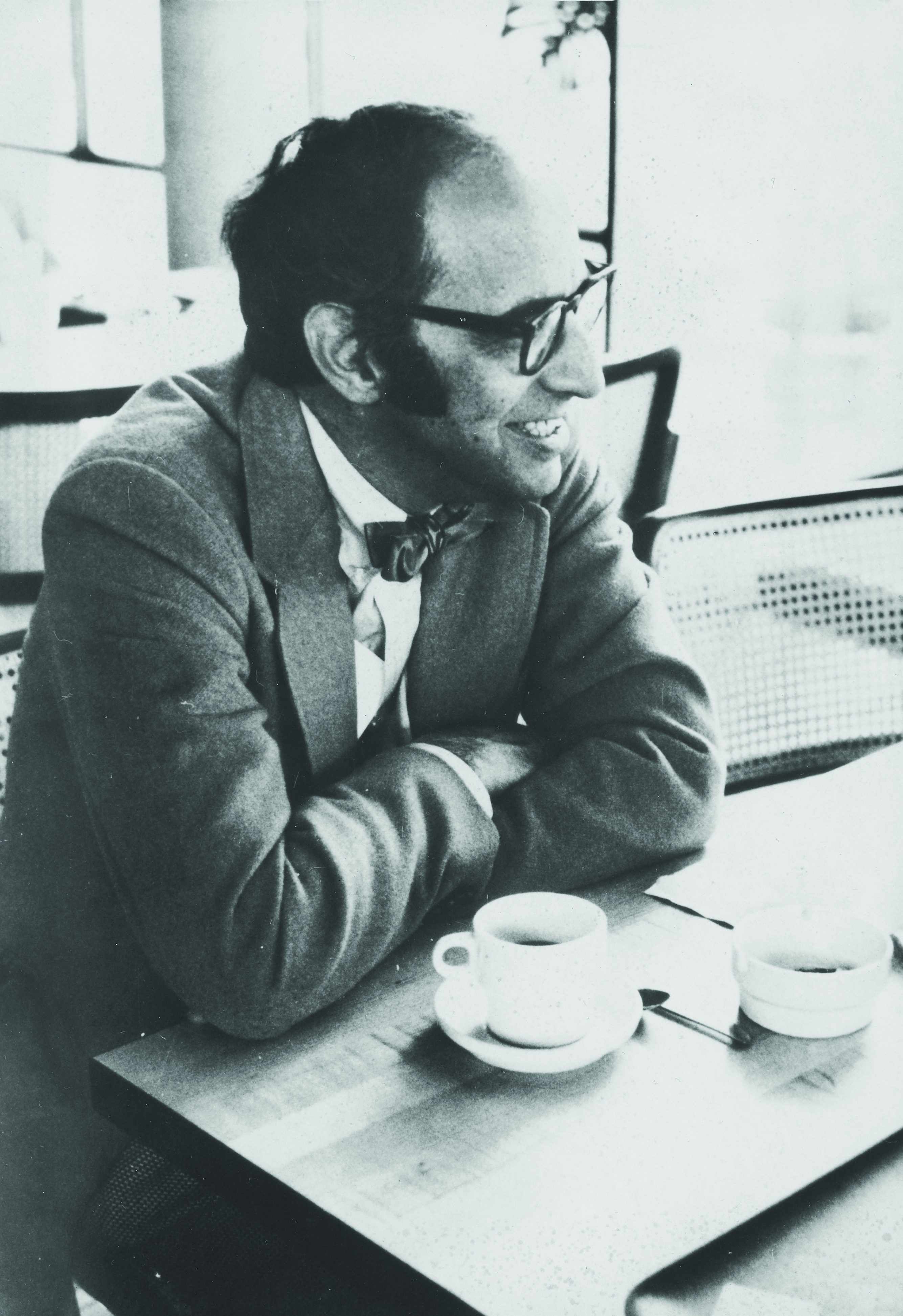 A middle-aged man wearing a suit and bow tie smiles with a cup of coffee in front of him.