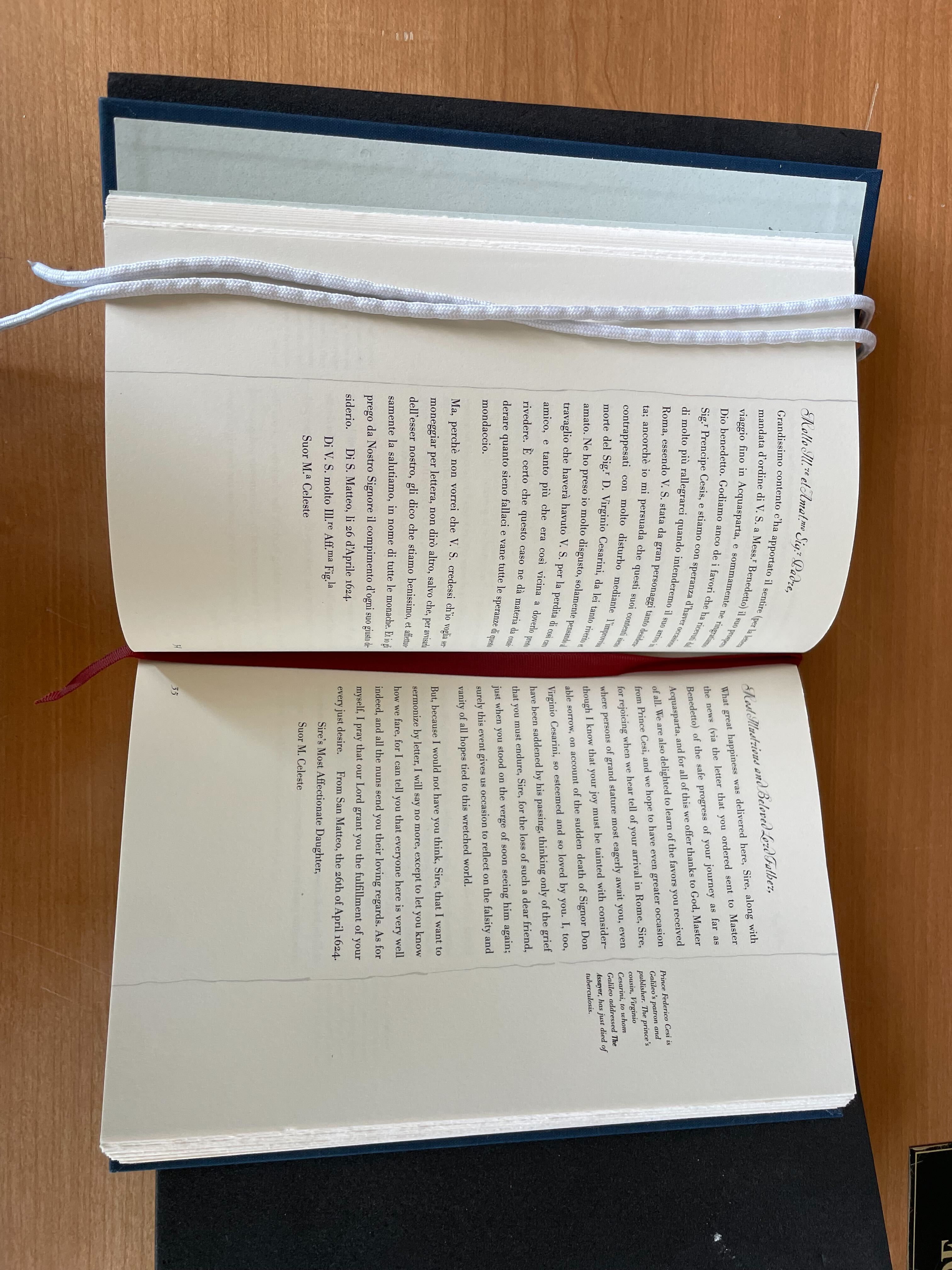 Book lying open on a table, with a red ribbon bookmark. Italian text on the left with English translation on the right.