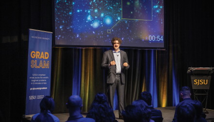 A tall man with long, wavy hair gives a presentation in front of a slide showing an image of globular clusters.