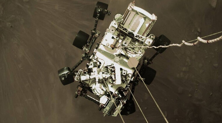 A still image showing the Perseverance rover approaching the surface of Mars, suspended by wires from a rocket-powered descent vehicle.
