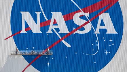 Workers suspended on a platform paint the NASA meatball logo on the side of a tall building.
