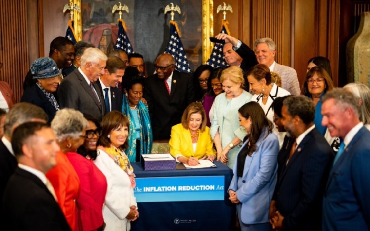 Rep. Pelosi sits at a desk surrounded by many House members as she signs the IRA