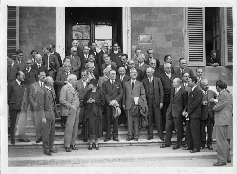 Black and white image of a crowd of well-dressed people standing outside of a building.