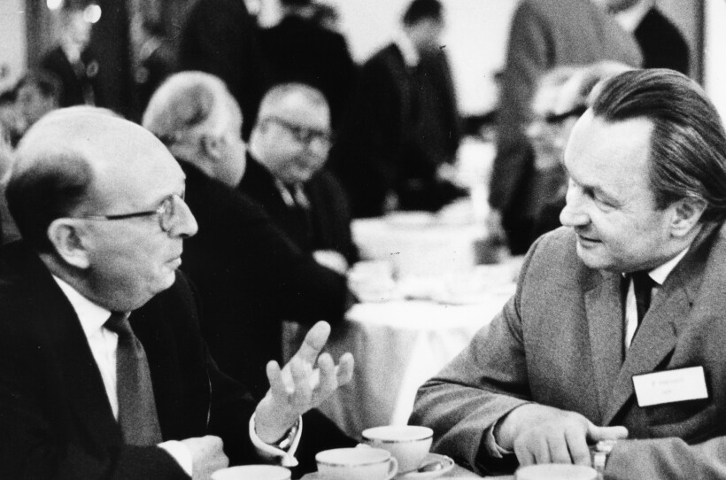 Black and white photo of two men sitting in a crowded room, looking at each other and conversing.
