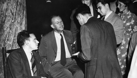 Five well-dressed men and one woman chat during a party. Morrison is one of two men seated at far left; the other stand.