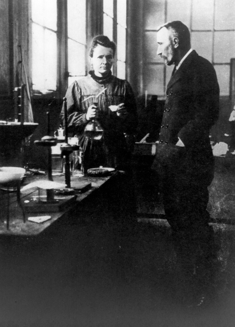 Marie and Pierre Curie stand at a workbench in their lab, Marie facing the camera and Pierre facing Marie.