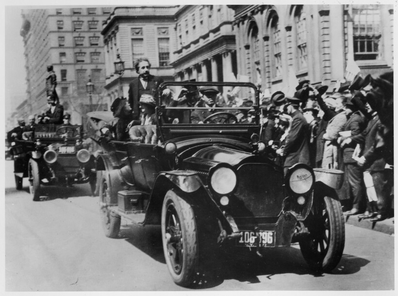 Albert Einstein stands in the back of a car in a motorcade down a New York City street lined with waving people.