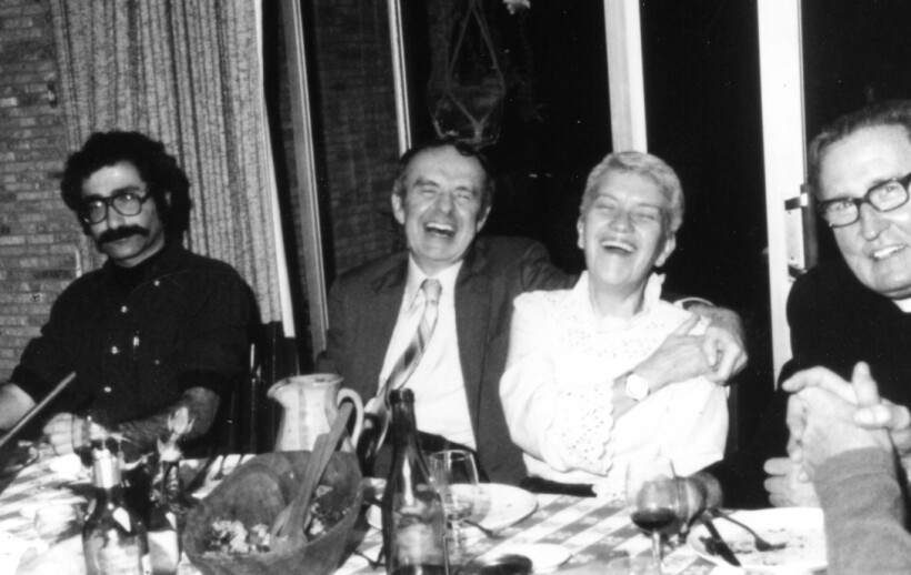 Jay Frogel, Allan Sandage, Vera Rubin, and Martin McCarthy sit in a row behind a table filled with food. Sandage has his arm around Rubin and they are both laughing.