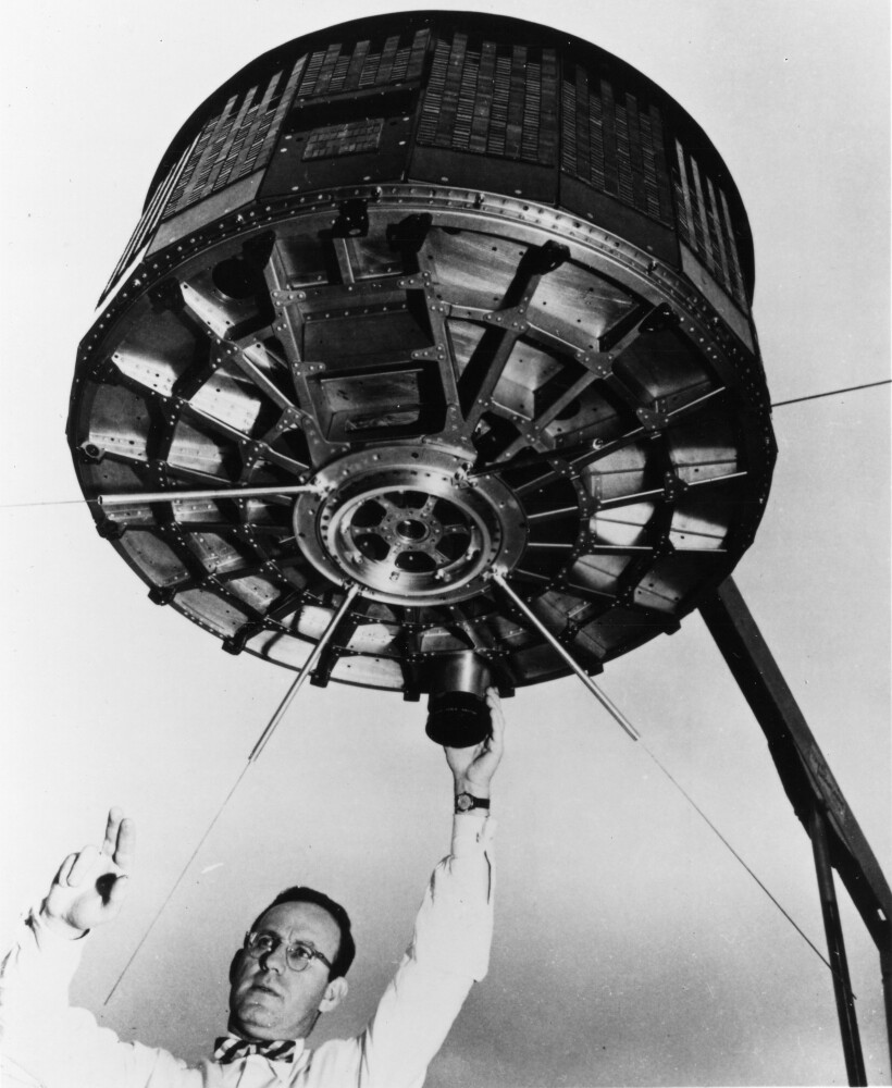 Man in a bow tie underneath an apparatus with one hand on the bottom of the apparatus and the other with two fingers in the air