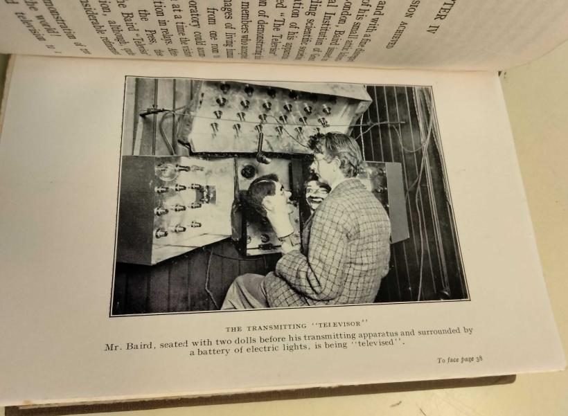 Image from book in black and white with a man seated in front of an apparatus, holding two doll heads in each hand, looking at one another