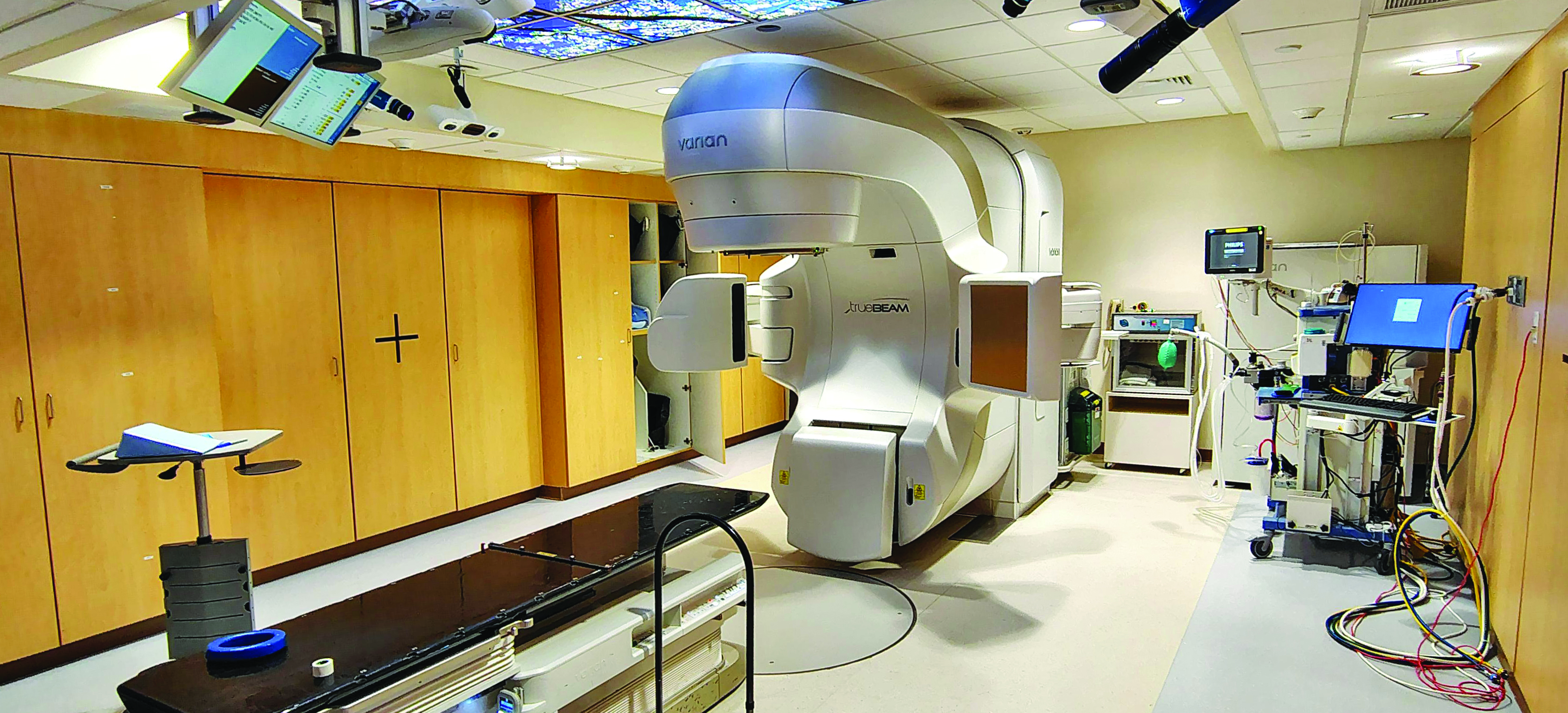 A room at a medical facility for conducting cancer radiation therapy. A table for a patient to lie down on is next to a large gray machine for administering radiation. There are cameras and screens mounted to the ceiling and a desk with a computer.